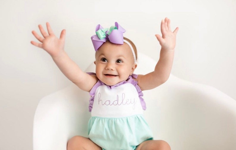 May include: A baby girl wearing a light green and white romper with a purple ruffle trim and the name "hadley" embroidered on the front. She is wearing a purple and green bow headband and has her arms raised in the air.