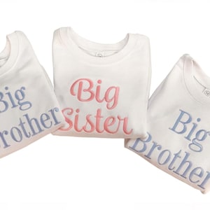 May include: Three white crew neck shirts with embroidered text. Two shirts say "Big Brother" in light blue, and one says "Big Sister" in pink. The shirts are folded and arranged on a white surface.