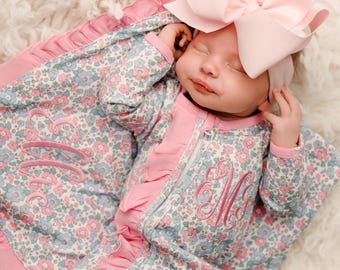 May include: A baby wearing a floral print onesie with pink trim and a matching pink bow headband. The onesie has a zipper closure and embroidered initial. The baby is lying on a fluffy white surface.