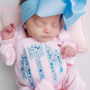 May include: A newborn baby wearing a light pink onesie with a floral monogram and a large light blue bow headband. The onesie has long sleeves and ruffled trim at the ankles.