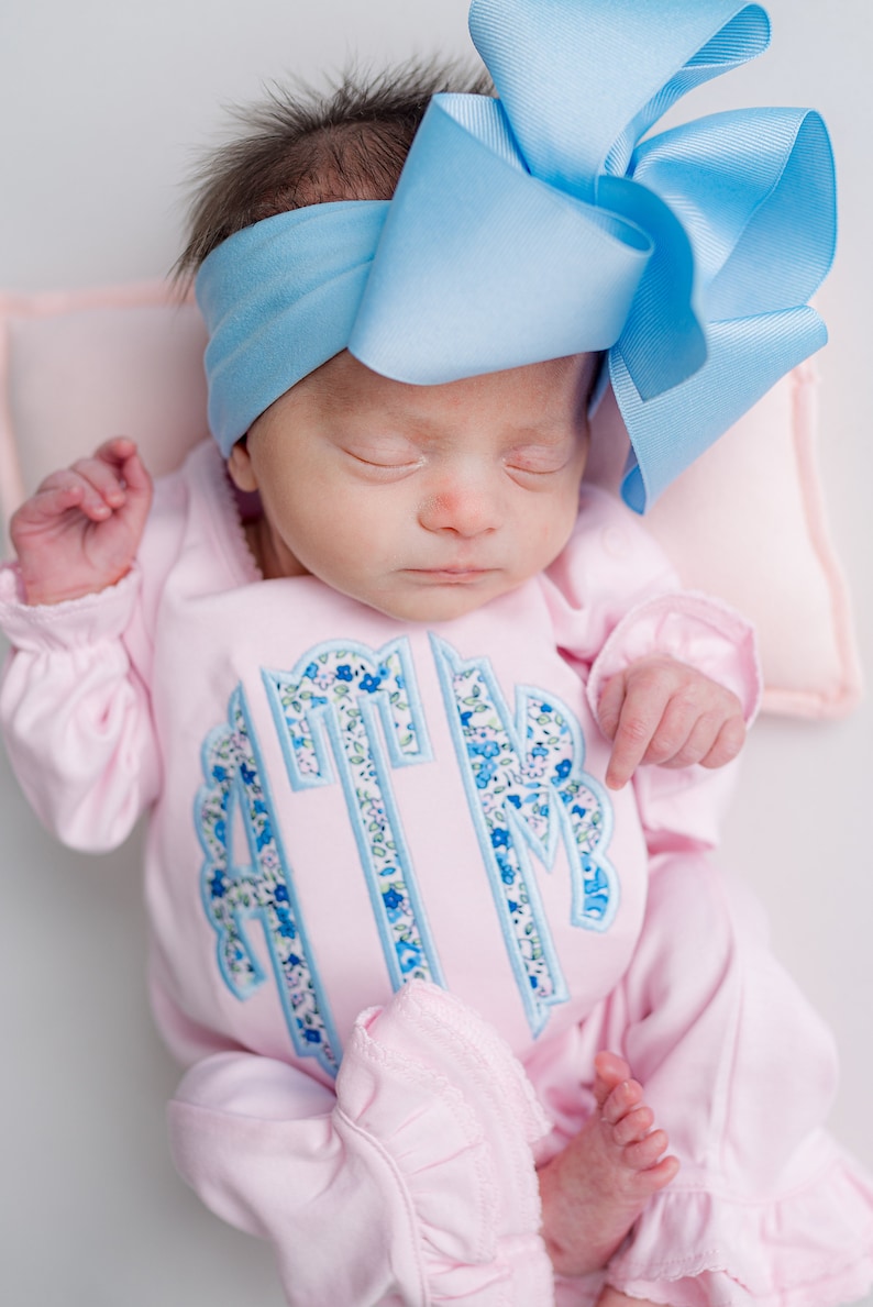 May include: A newborn baby wearing a light pink onesie with a floral monogram and a large light blue bow headband. The onesie has long sleeves and ruffled trim at the ankles.