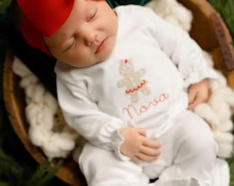 May include: A newborn baby wearing a white onesie with a gingerbread man design and the name "Nova" embroidered in red. The baby is nestled in a wooden bowl surrounded by greenery and a white blanket, with a large red bow on their head.