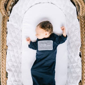 May include: A newborn baby boy wearing a navy blue gown with a gray embroidered name tag that says "William" is sleeping in a white wicker bassinet.