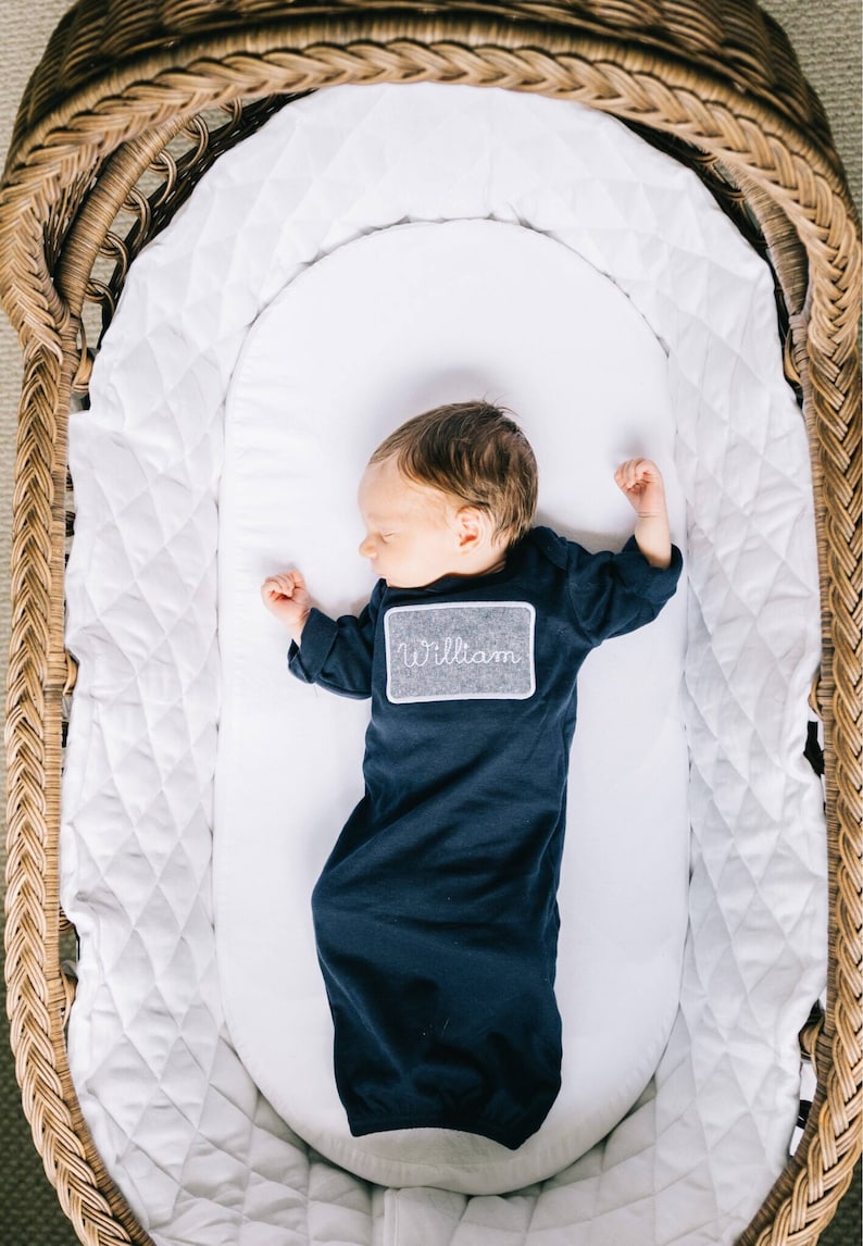 May include: A newborn baby boy wearing a navy blue gown with a gray embroidered name tag that says "William" is sleeping in a white wicker bassinet.