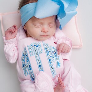 May include: A newborn baby wearing a light pink onesie with a floral monogram and ruffled trim. The baby is also wearing a large, light blue bow headband. The baby is lying on a light pink pillow.