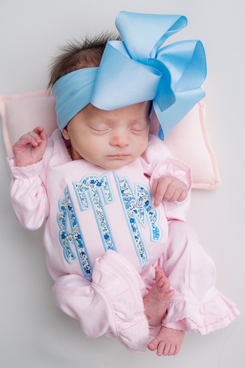 May include: A newborn baby wearing a light pink onesie with a floral monogram and ruffled trim. The baby is also wearing a large, light blue bow headband. The baby is lying on a light pink pillow.