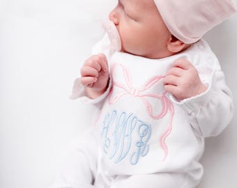 Puede incluir: Un pelele blanco para bebé con un lazo y un monograma bordados en rosa y azul. El bebé lleva una diadema rosa con un lazo grande.