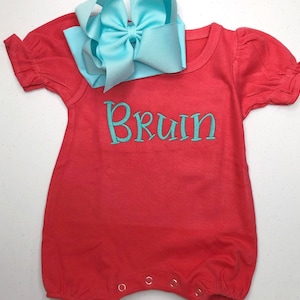 May include: A coral red baby onesie with short sleeves and a turquoise blue bow on top. The onesie has the name "Bruin" embroidered on the front in turquoise blue thread.