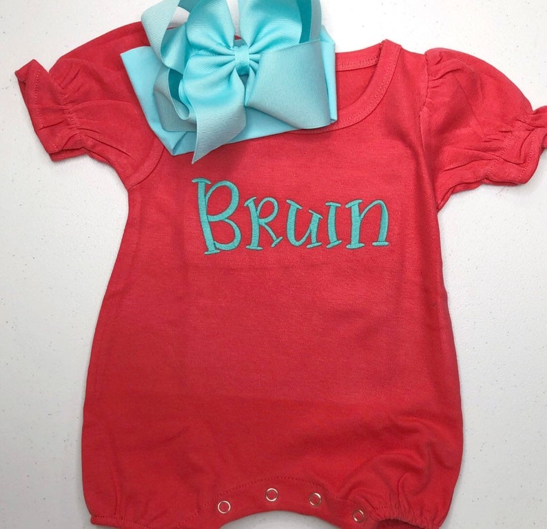 May include: A coral red baby onesie with short sleeves and a turquoise blue bow on top. The onesie has the name "Bruin" embroidered on the front in turquoise blue thread.
