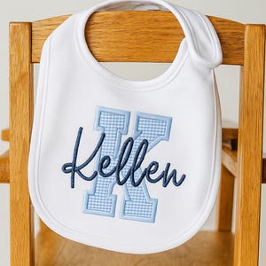 May include: White baby bib with a blue gingham letter "K" and the name "Kellen" embroidered in a cursive font. The bib is hanging on a wooden chair. A perfect gift for a baby shower.