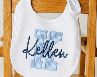 May include: A white baby bib featuring a large light blue gingham block letter K embroidered in the center. The name Kellen is stitched over the letter in a dark navy blue cursive script. The bib has a rounded shape with a side closure.