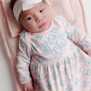 Pu&ograve; includere: Un bambino con una fascia bianca e un grande fiocco bianco. Il bambino indossa un vestito con stampa floreale rosa chiaro e blu con un monogramma. Il bambino &egrave; sdraiato su una coperta rosa chiaro.
