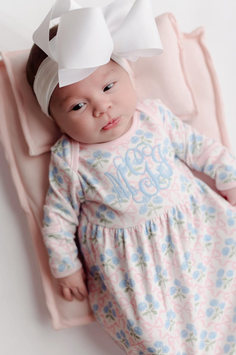 Pu&ograve; includere: Un bambino con una fascia bianca e un grande fiocco bianco. Il bambino indossa un vestito con stampa floreale rosa chiaro e blu con un monogramma. Il bambino &egrave; sdraiato su una coperta rosa chiaro.
