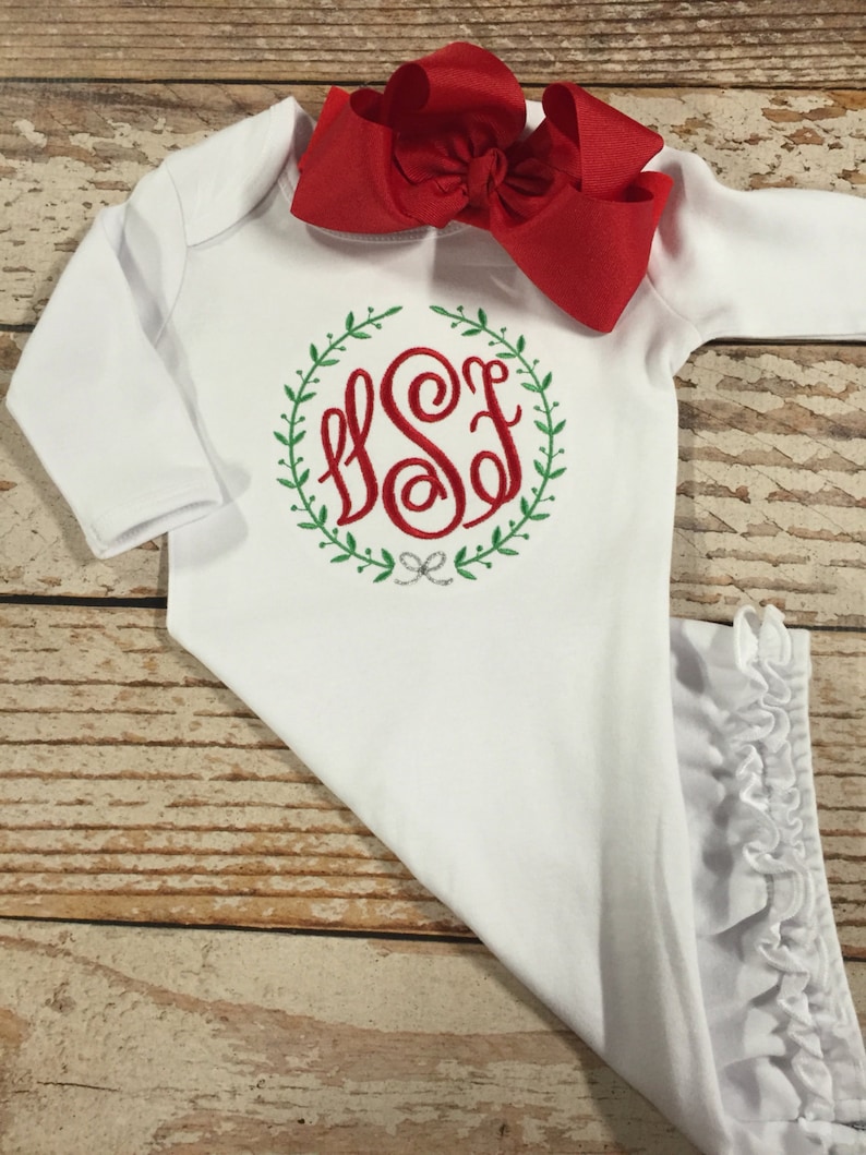 Pode incluir: Batista branca para beb&eacute; com mangas compridas e uma bainha com babados. A bata tem um monograma vermelho com as letras "SSF" dentro de uma coroa verde.