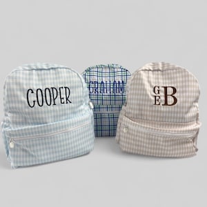 May include: Three small backpacks with gingham patterns. One is light blue with "COOPER" embroidered in black. Another is blue and green with "GRAHAM" embroidered in blue. The third is tan with "GEB" embroidered in brown.