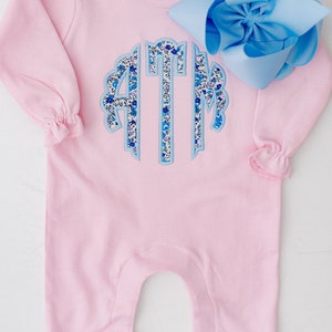 May include: A light pink baby romper with long sleeves and ruffled cuffs and ankles. The romper features a floral monogram in blue and white. A large light blue bow is placed on the side.