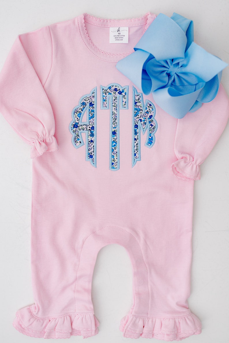 May include: A light pink baby romper with long sleeves and ruffled cuffs and ankles. The romper features a floral monogram in blue and white. A large light blue bow is placed on the side.