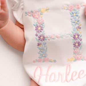 May include: White baby onesie with a floral embroidered letter "H" in pastel colors. The name "Harlee" is embroidered in pink script below the letter. The onesie has ruffled sleeves.