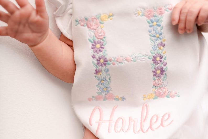 May include: White baby onesie with a floral embroidered letter "H" in pastel colors. The name "Harlee" is embroidered in pink script below the letter. The onesie has ruffled sleeves.