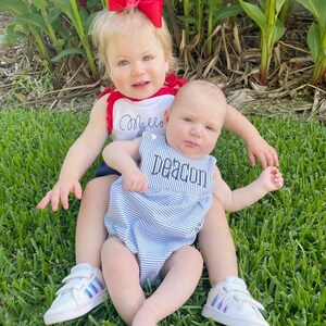 May include: Two young children are sitting on green grass. One child wears a red and white top with the name "Mello" embroidered on it and a red bow. The other child wears a blue and white striped romper with the name "Deacon" embroidered on it. Both children are wearing white sneakers.