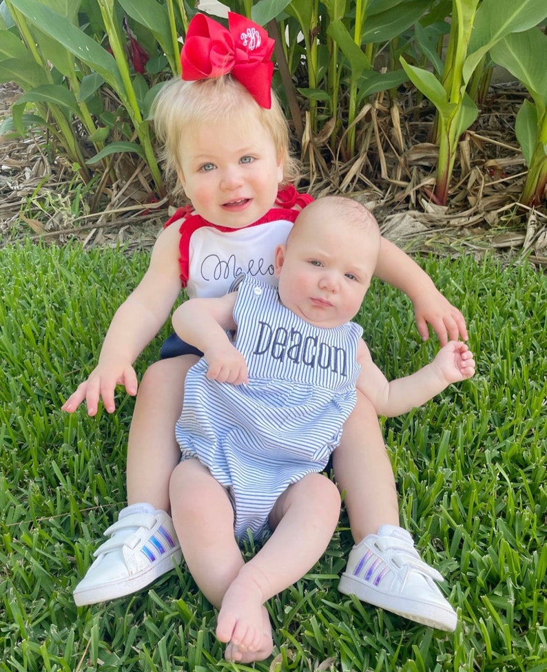 May include: Two young children are sitting on green grass. One child wears a red and white top with the name "Mello" embroidered on it and a red bow. The other child wears a blue and white striped romper with the name "Deacon" embroidered on it. Both children are wearing white sneakers.