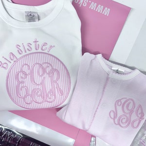 May include: Two pink and white striped shirts with embroidered monograms. The larger shirt says "Big Sister" and has the monogram "EGR". The smaller shirt has the monogram "SG".