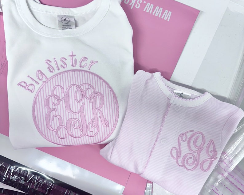 May include: Two pink and white striped shirts with embroidered monograms. The larger shirt says "Big Sister" and has the monogram "EGR". The smaller shirt has the monogram "SG".