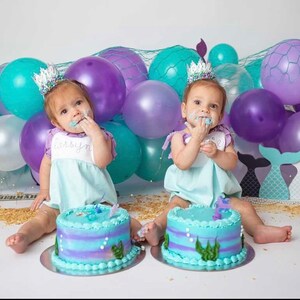 May include: Two babies wearing turquoise and white dresses with mermaid crowns are sitting in front of a backdrop of purple and turquoise balloons and a net. They are each eating a blue and purple cake with a mermaid tail on top.
