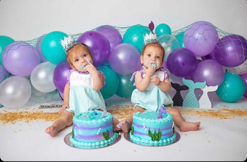 May include: Two babies wearing turquoise and white dresses with mermaid crowns are sitting in front of a backdrop of purple and turquoise balloons and a net. They are each eating a blue and purple cake with a mermaid tail on top.