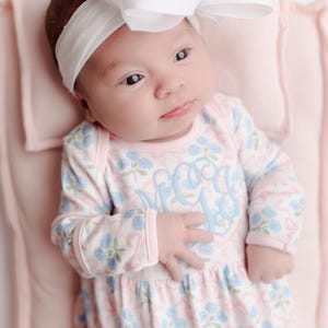 Pu&ograve; includere: Un bambino che indossa una fascia bianca con un grande fiocco e un body a fantasia floreale. Il body presenta un monogramma azzurro e un design floreale rosa e blu. Il bambino &egrave; sdraiato su una coperta rosa.