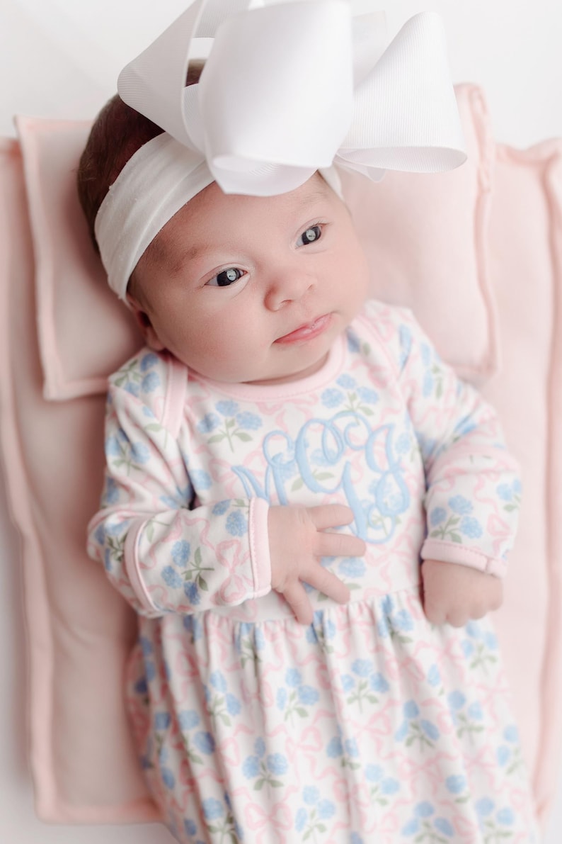 Pu&ograve; includere: Un bambino che indossa una fascia bianca con un grande fiocco e un body a fantasia floreale. Il body presenta un monogramma azzurro e un design floreale rosa e blu. Il bambino &egrave; sdraiato su una coperta rosa.