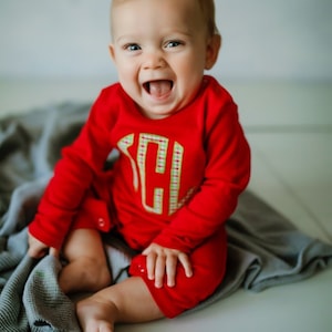 May include: A baby in a red long-sleeved onesie with a green and white plaid monogrammed letter on the front is sitting on a gray blanket. The baby is smiling and has a happy expression.