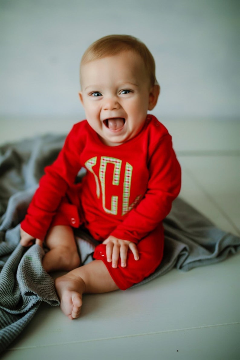 May include: A baby in a red long-sleeved onesie with a green and white plaid monogrammed letter on the front is sitting on a gray blanket. The baby is smiling and has a happy expression.