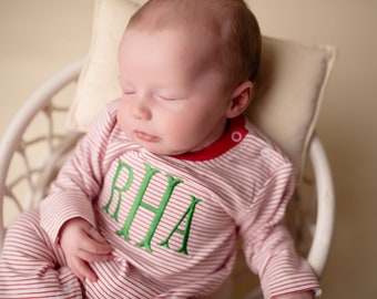 May include: A baby wearing a red and white striped sleeper with green embroidered letters that spell "RHA". The sleeper has a red collar and a red button on the shoulder.