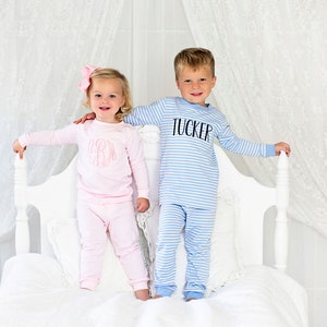 Puede incluir: Dos ni&ntilde;os con pijamas a rayas a juego. La ni&ntilde;a lleva un pijama rosa con un monograma en la parte delantera. El ni&ntilde;o lleva un pijama azul con el nombre "Tucker" en la parte delantera. Est&aacute;n de pie en una cama blanca.