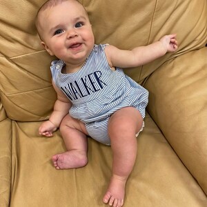 May include: A smiling baby wearing a blue and white striped romper with the name "WALKER" embroidered on the front. The baby is sitting on a tan leather couch, with arms outstretched.