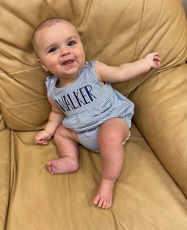 May include: A smiling baby wearing a blue and white striped romper with the name "WALKER" embroidered on the front. The baby is sitting on a tan leather couch, with arms outstretched.