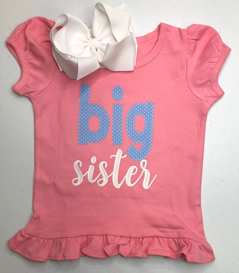 K&ouml;nnte beinhalten: Rosa T-Shirt mit kurzen &Auml;rmeln und R&uuml;schen am Saum. Das Shirt hat ein blaues und wei&szlig;es Tupfenmuster mit der Aufschrift "gro&szlig;e Schwester".