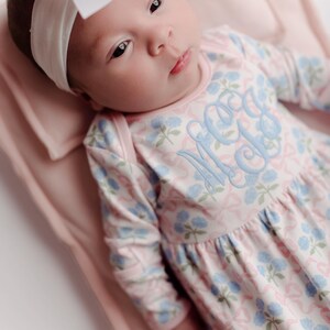 Pu&ograve; includere: Un bambino che indossa un vestito floreale rosa e blu con una fascia abbinata e un grande fiocco bianco. L'abito presenta un monogramma azzurro. Il bambino &egrave; sdraiato su una coperta rosa.