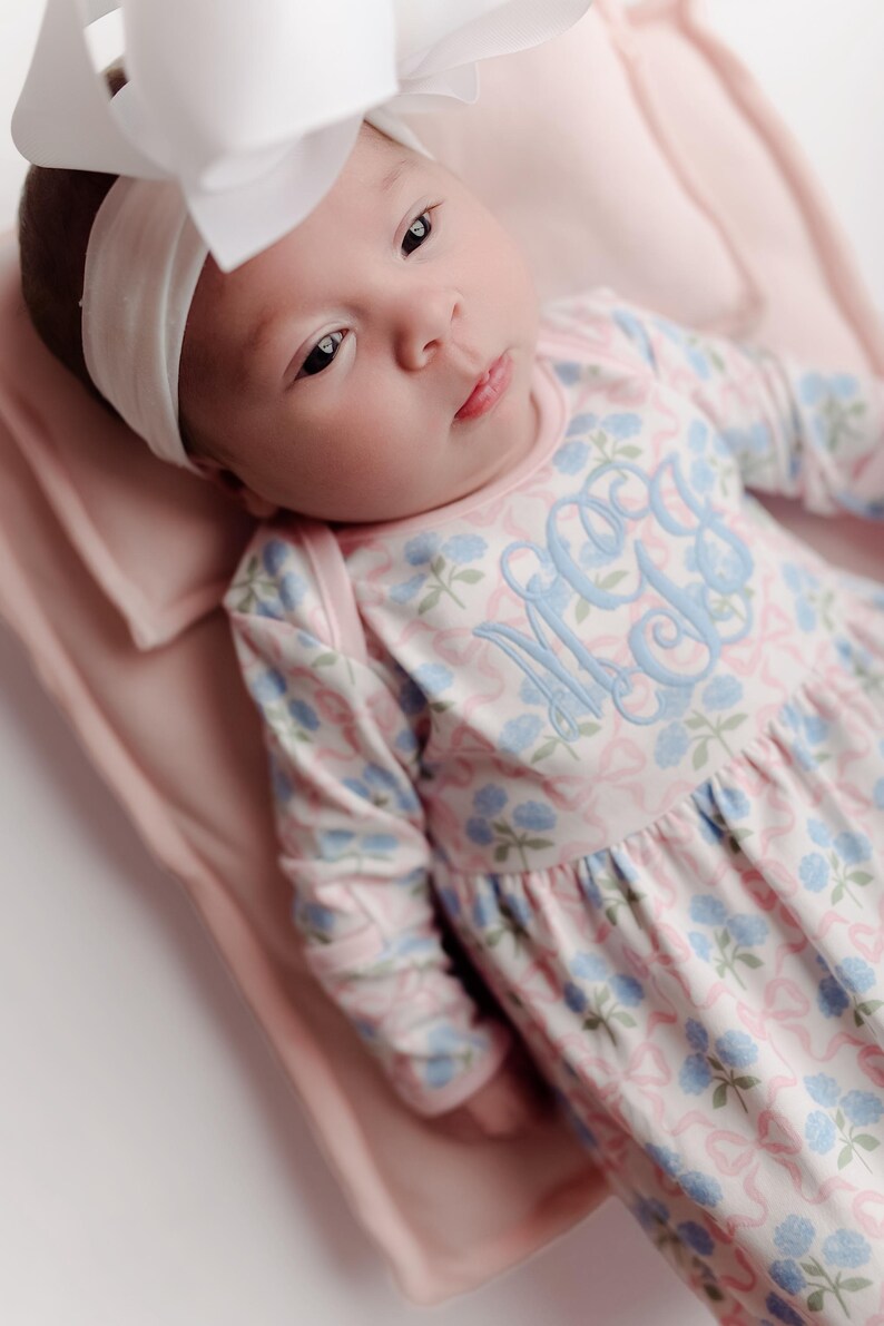 Pu&ograve; includere: Un bambino che indossa un vestito floreale rosa e blu con una fascia abbinata e un grande fiocco bianco. L'abito presenta un monogramma azzurro. Il bambino &egrave; sdraiato su una coperta rosa.