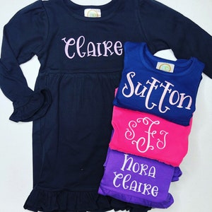 May include: A stack of four navy blue, pink, and purple long-sleeved shirts with embroidered names, including "Claire", "Sutton", "Nora", and "SFJ".