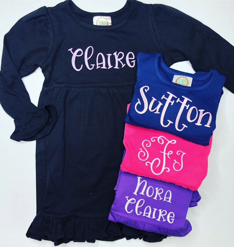 May include: A stack of four navy blue, pink, and purple long-sleeved shirts with embroidered names, including "Claire", "Sutton", "Nora", and "SFJ".