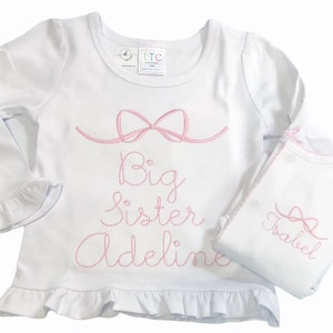 May include: White long-sleeved shirt with a pink bow and the text "Big Sister Adeline" embroidered on the front. A white onesie with a pink bow and the text "Isabel" embroidered on the front is also shown.