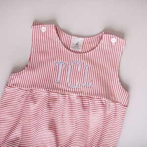 May include: Red and white striped romper with light blue embroidered initials. The sleeveless garment has a gathered waist and button closures on the shoulders. The fabric is a soft knit, suitable for a baby or toddler.
