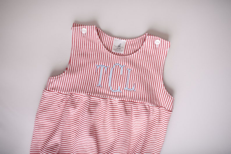 May include: Red and white striped romper with light blue embroidered initials. The sleeveless garment has a gathered waist and button closures on the shoulders. The fabric is a soft knit, suitable for a baby or toddler.