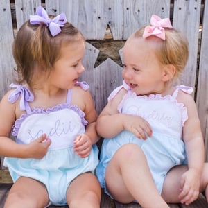 May include: Two little girls wearing matching light blue and white dresses with their names embroidered on the front. The girl on the left has a purple bow in her hair and her name is Sadie. The girl on the right has a pink bow in her hair and her name is Audrey.
