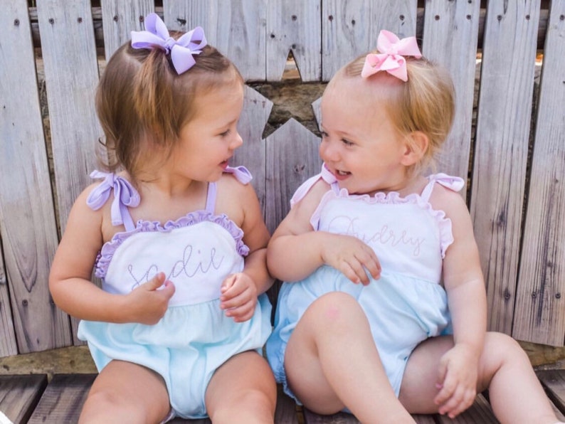 May include: Two little girls wearing matching light blue and white dresses with their names embroidered on the front. The girl on the left has a purple bow in her hair and her name is Sadie. The girl on the right has a pink bow in her hair and her name is Audrey.