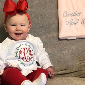 Pode incluir: Uma menina pequena vestindo uma camisa branca de manga comprida com um monograma vermelho e folhas verdes, cal&ccedil;as vermelhas e um la&ccedil;o vermelho no cabelo. A beb&ecirc; est&aacute; sentada em um sof&aacute; cinza com um cobertor rosa atr&aacute;s dela. O cobertor tem o texto "Caroline C Abril 8" bordado.