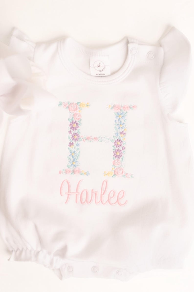 May include: White baby romper with floral embroidered letter "H" and the name "Harlee" in pink script. The letter is filled with pastel flowers. The romper has short sleeves with ruffled edges and snap closures.