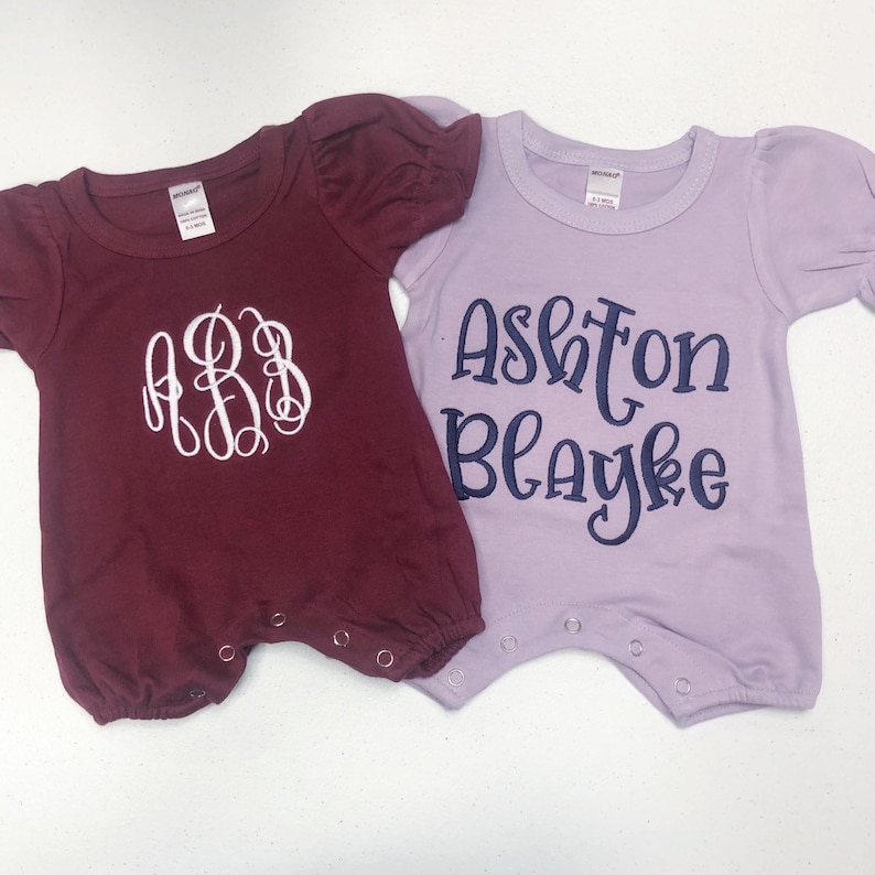 May include: Two baby onesies, one burgundy with white embroidered initials "ABB" and one lavender with navy blue embroidered text "Ashton Blayke".